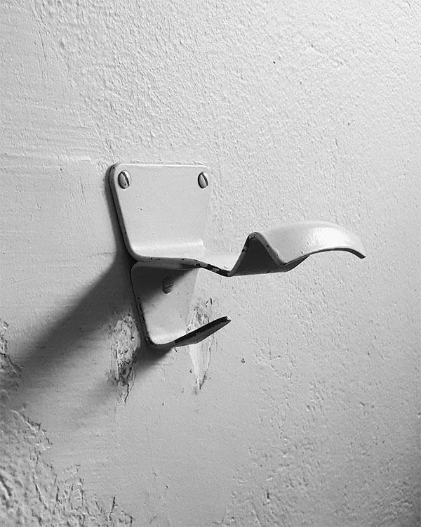 A double hook made of metal, gray paint, screwed to a wall. Found at Piscine Municipale de Apt in Apt, France, 2018.