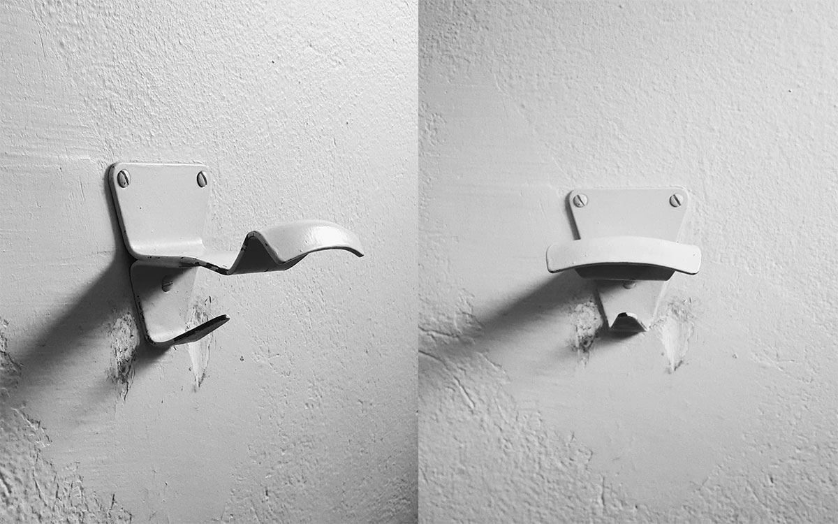 A double hook made of metal, gray paint, screwed to a wall. Found at Piscine Municipale de Apt in Apt, France, 2018.